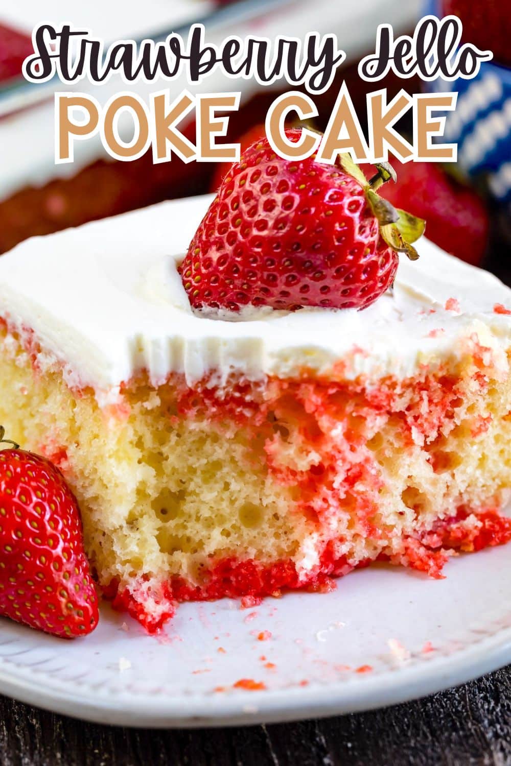 A slice of yellow poke cake with white frosting and red strawberry filling is topped with a fresh strawberry. Another strawberry sits on the plate next to the cake. Bold text reads “Strawberry Jello Poke Cake.”.
