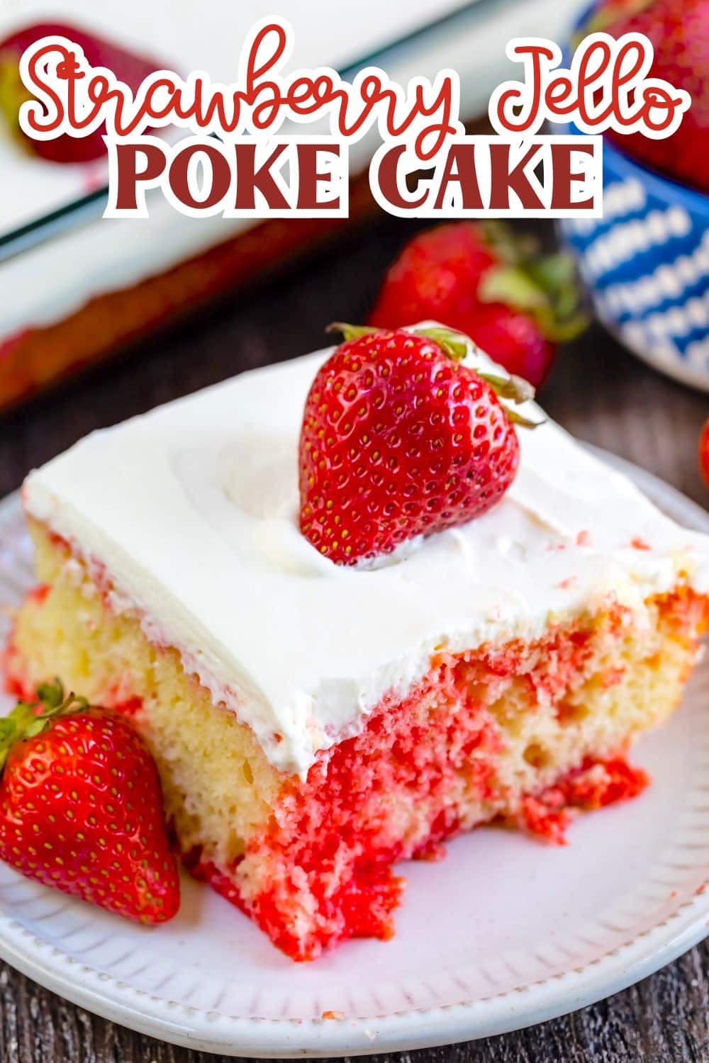 A slice of strawberry jello poke cake topped with whipped cream and a fresh strawberry, served on a white plate with additional strawberries around it. The cake has visible red jello sections.