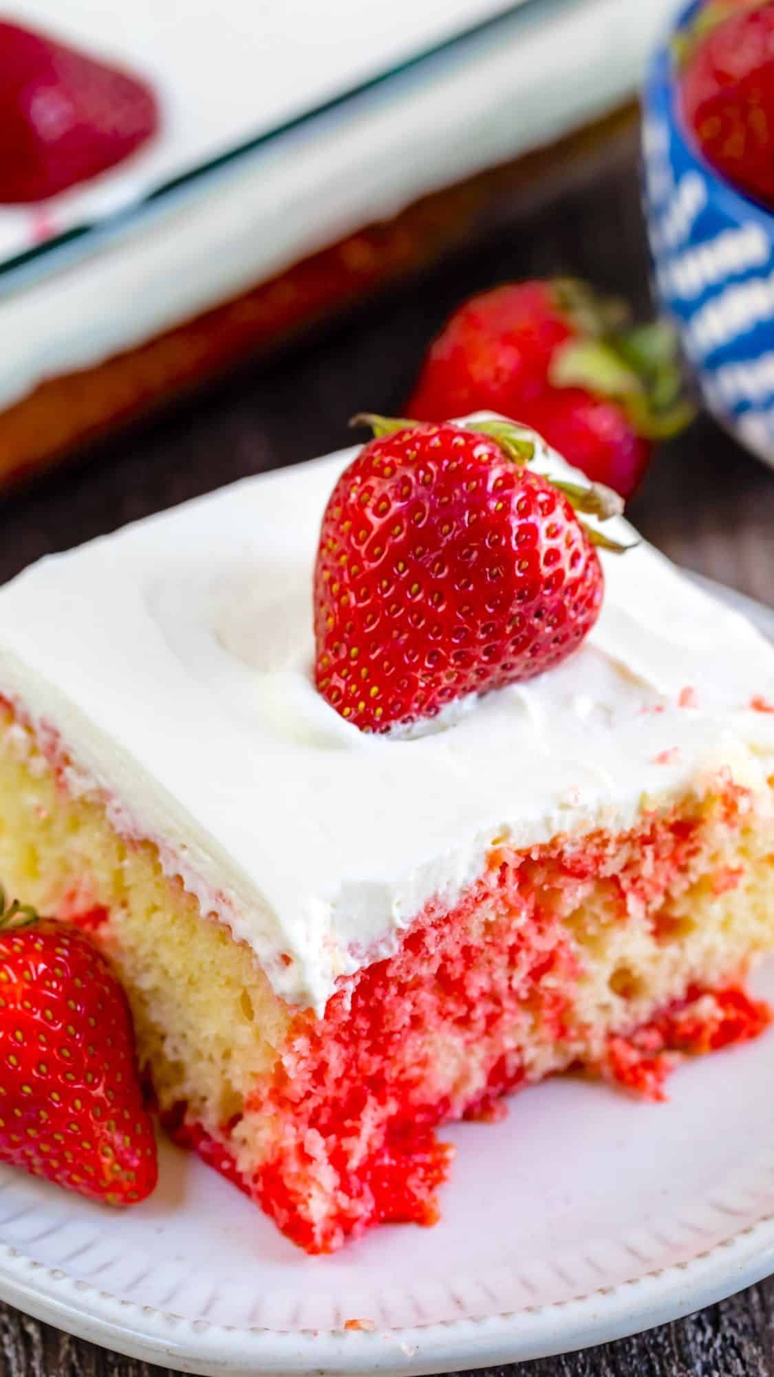 A slice of strawberry poke cake with white frosting and a whole strawberry on top, sitting on a white plate. The cake has visible red strawberry gelatin throughout and fresh strawberries beside it.