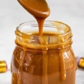 A spoon drips thick caramel sauce into a glass jar, with caramel running down the sides. Two unwrapped Werthers Original candies are placed next to the jar on a light surface.