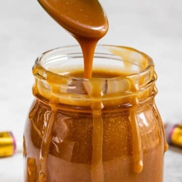 A spoon drips thick caramel sauce into a glass jar, with caramel running down the sides. Two unwrapped Werthers Original candies are placed next to the jar on a light surface.