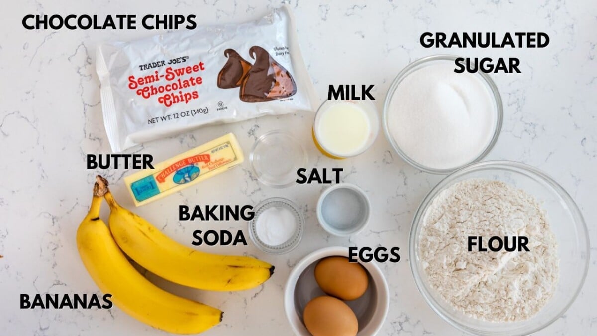 Ingredients for baking are arranged on a white surface, including bananas, a stick of butter, chocolate chips, baking soda, salt, eggs, milk, granulated sugar, and a bowl of flour. Each item is labeled in bold text.