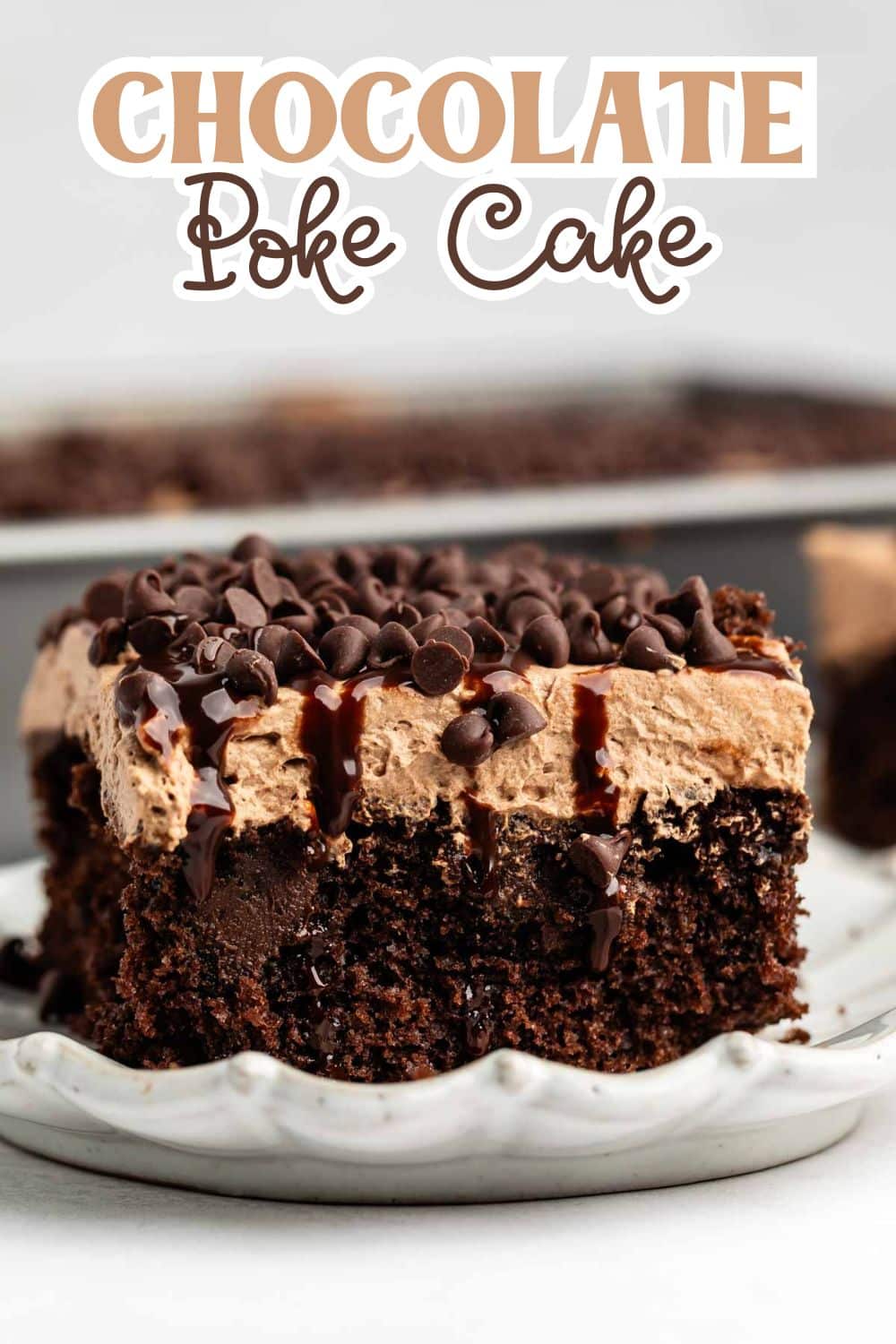 A slice of chocolate poke cake topped with fluffy chocolate frosting, chocolate chips, and chocolate drizzle sits on a white scalloped plate; more cake is visible in the background. Text reads “Chocolate Poke Cake.”.