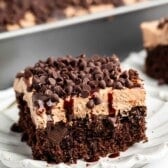 A square slice of chocolate cake topped with chocolate frosting, mini chocolate chips, and chocolate drizzle sits on a white plate, with a pan of more cake in the background.