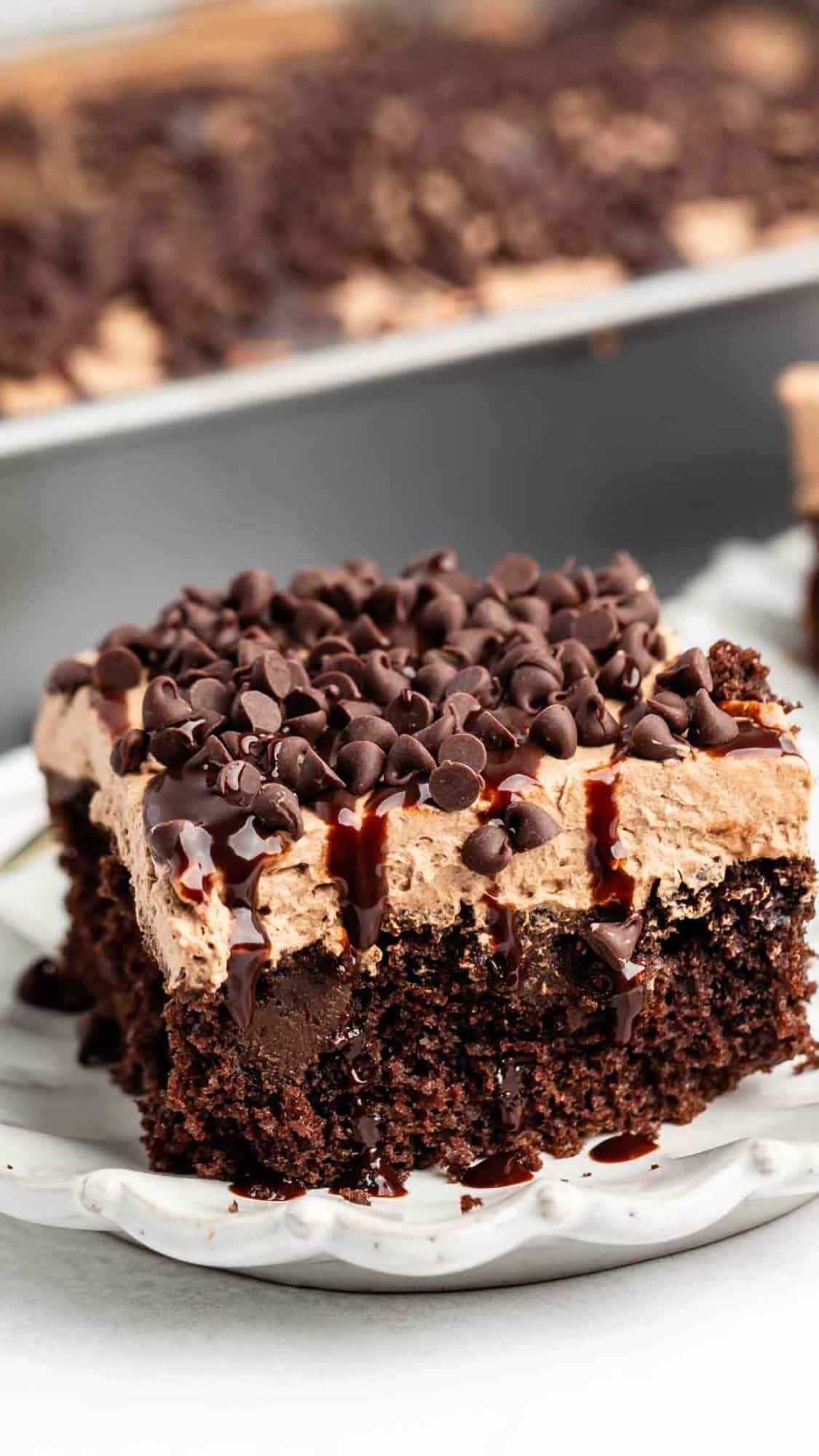 A close-up of a slice of chocolate cake topped with chocolate mousse, chocolate chips, and a drizzle of chocolate sauce, served on a white plate. The rest of the cake is visible in the background.