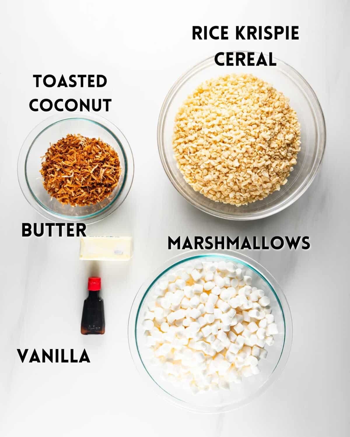 Top-down view of ingredients for a recipe: a bowl of Rice Krispie cereal, a bowl of mini marshmallows, a bowl of toasted coconut, a stick of butter, and a bottle of vanilla extract, labeled accordingly.