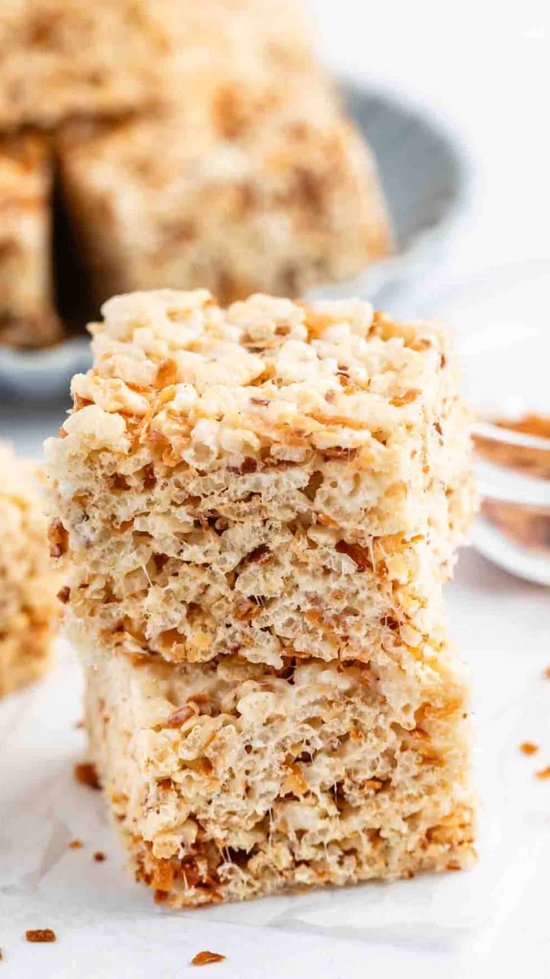 A close-up of two stacked toasted coconut Rice Krispie treats, showing their crispy, golden texture. More treats and toasted coconut flakes are visible in the blurred background.