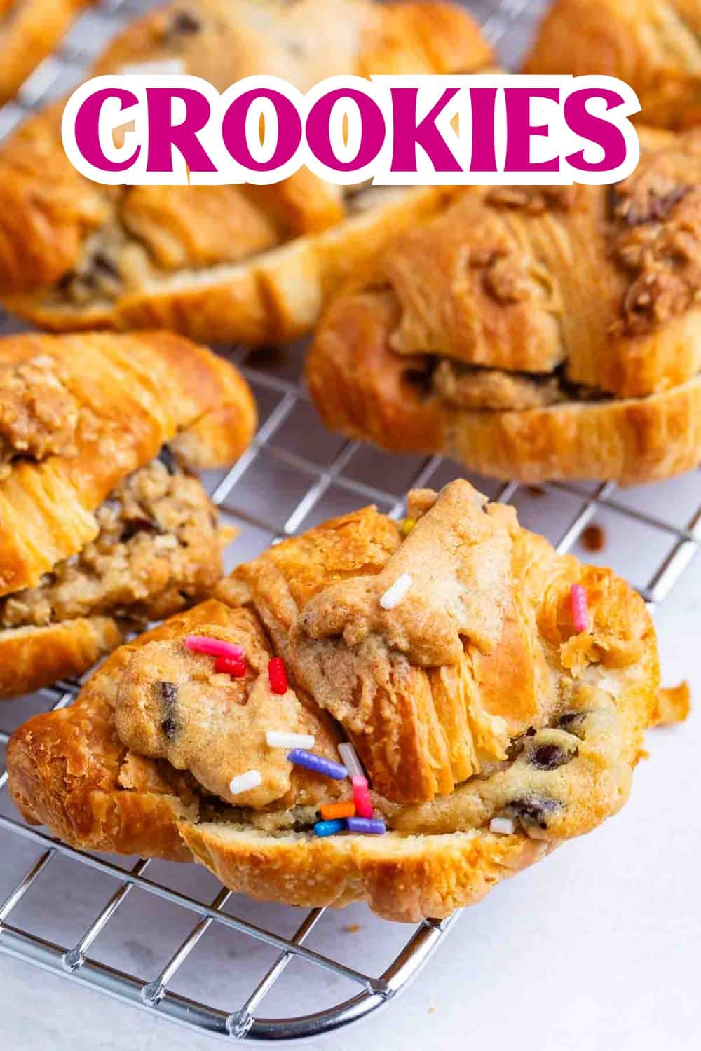 A wire rack holds several croissants stuffed with chocolate chip cookie dough and topped with colorful sprinkles. Bold pink and white text above reads CROOKIES.