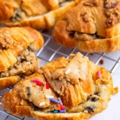 Freshly baked croissants stuffed with chocolate chip cookie dough, some topped with colorful sprinkles, are cooling on a wire rack. The pastries are golden brown and flaky.
