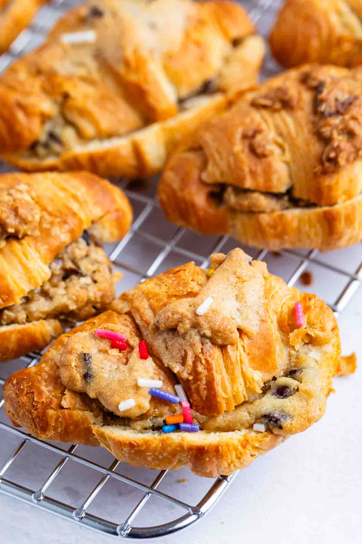 Freshly baked croissants stuffed with chocolate chip cookie dough, some topped with colorful sprinkles, are cooling on a wire rack. The pastries are golden brown and flaky.