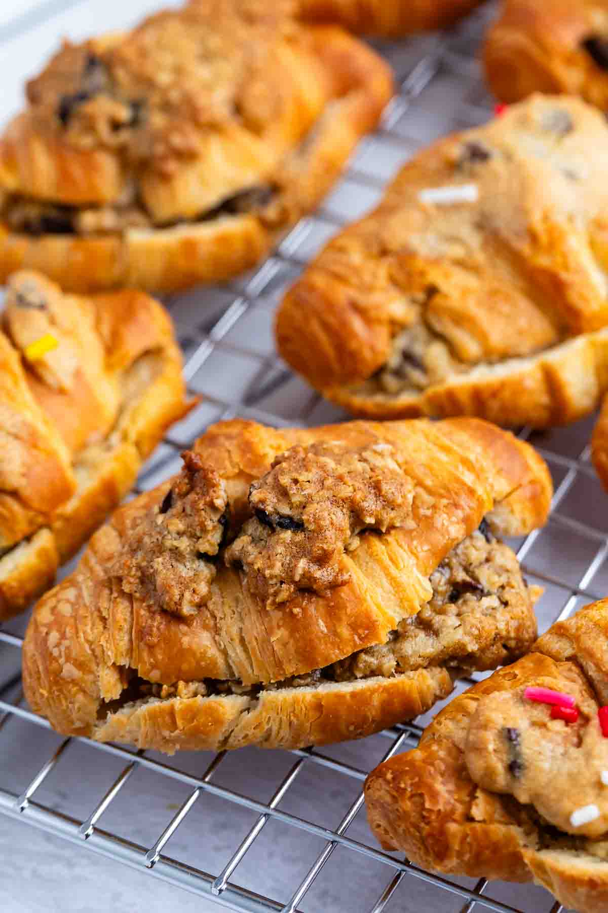 Golden-brown croissants filled with a crumbly, streusel-like topping rest on a wire cooling rack. The croissants appear flaky and freshly baked, with textured, sweet fillings visible inside.