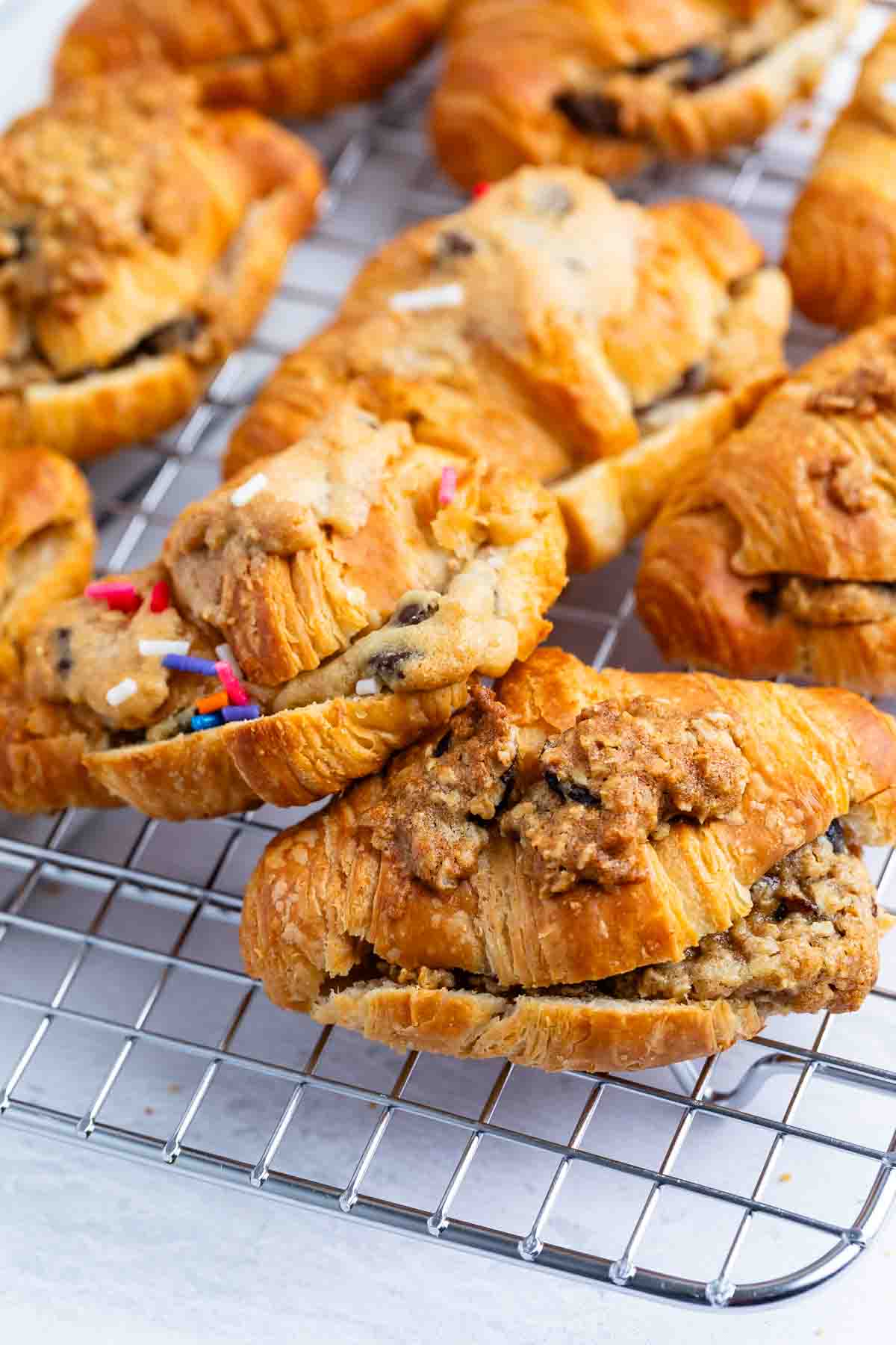 Several croissants filled with cookie dough and topped with colorful sprinkles are arranged on a wire cooling rack. The croissants have a flaky golden crust and chunky cookie dough filling.