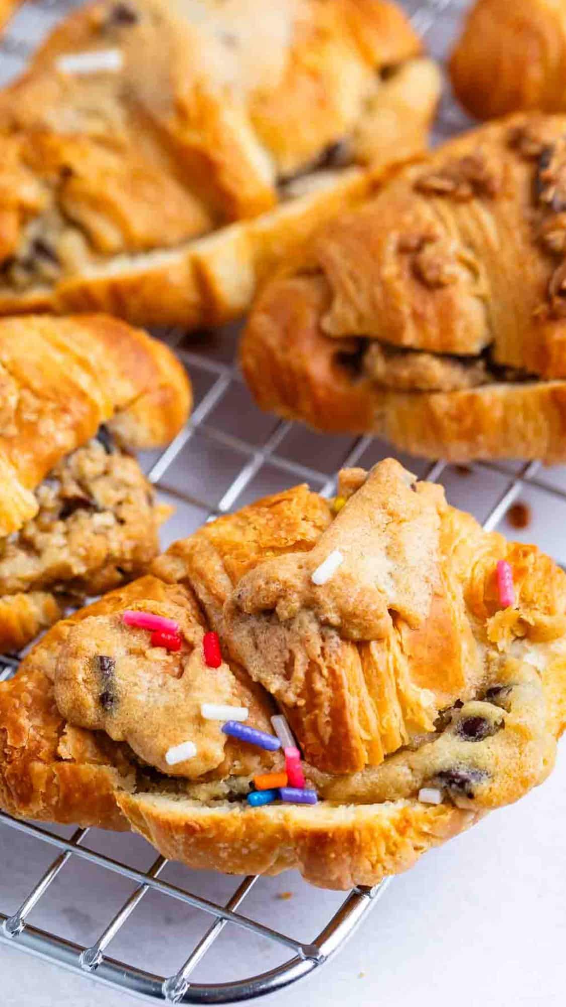 Golden croissants filled with cookie dough and chocolate chips, some topped with colorful sprinkles, cooling on a wire rack.