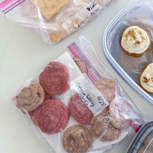 Three containers with baked goods: a bag of rectangular bars, a bag of strawberry cookies on paper towels, and a plastic box with cupcakes topped with icing, all labeled and stored for freshness.