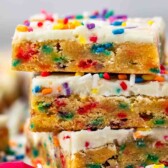A close-up of three stacked, frosted blondie bars filled with colorful rainbow sprinkles. The bars have a soft, chewy texture and are topped with white icing and extra sprinkles.