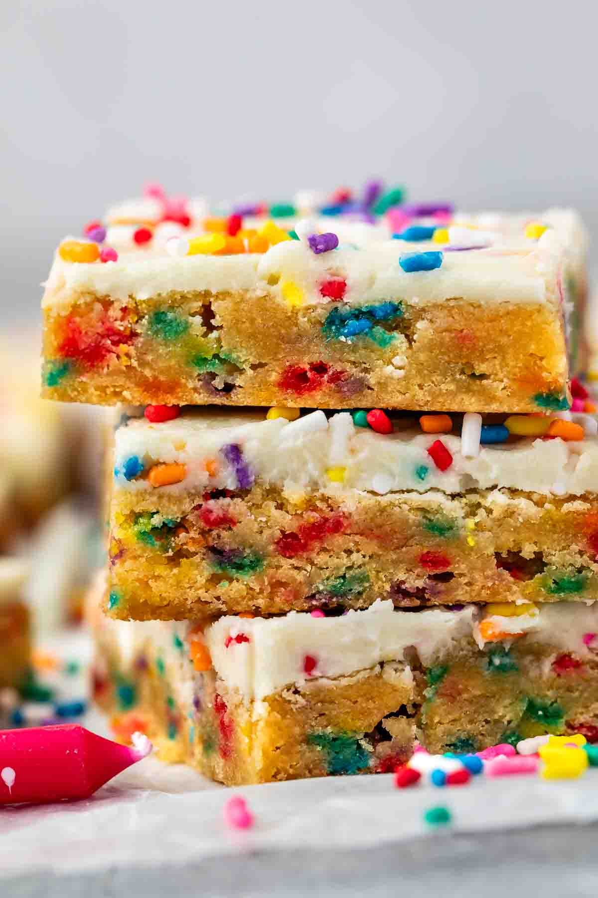 A close-up of three stacked, frosted blondie bars filled with colorful rainbow sprinkles. The bars have a soft, chewy texture and are topped with white icing and extra sprinkles.