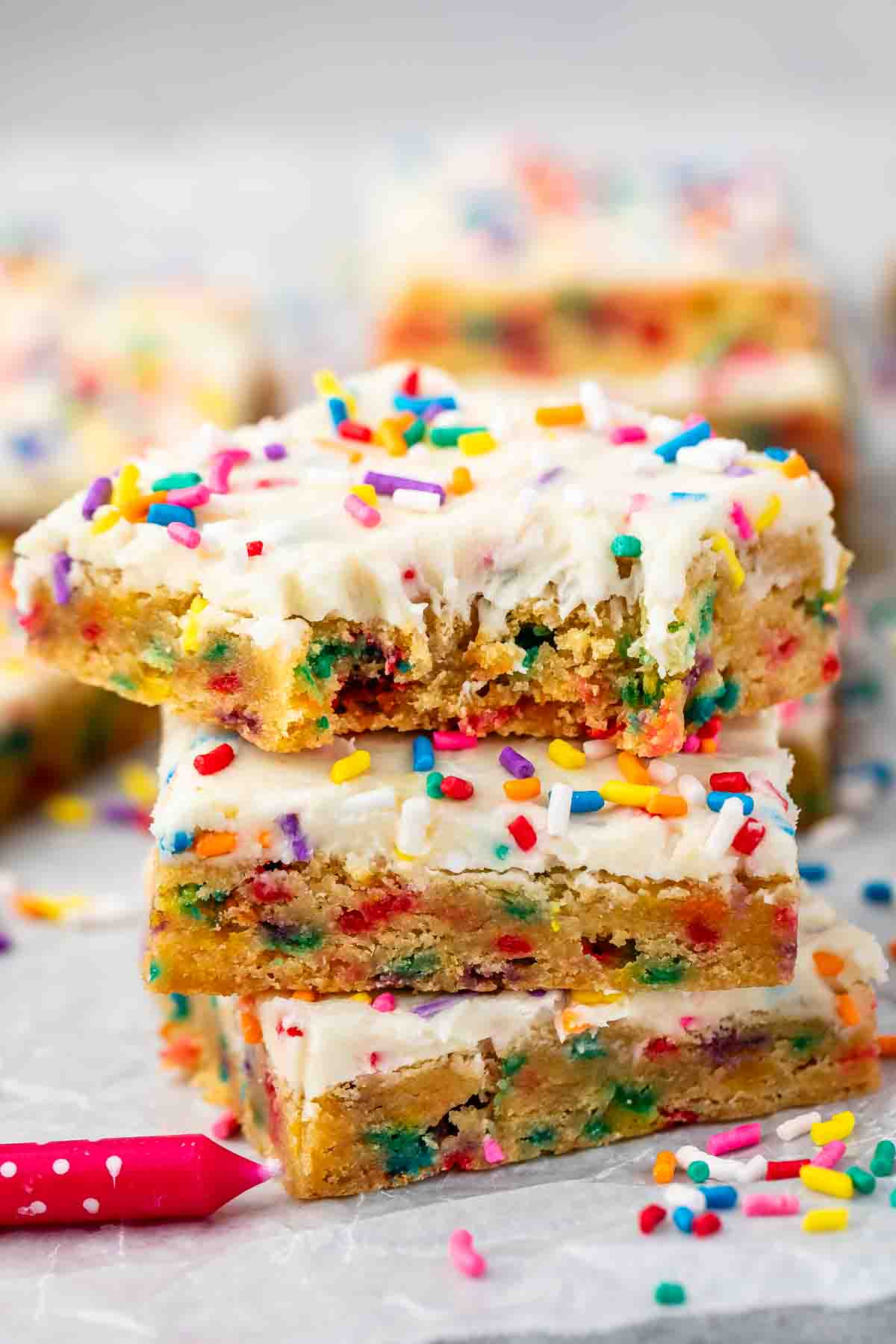 Three frosted funfetti cake bars stacked on top of each other, topped with colorful rainbow sprinkles. The top bar has a bite taken out of it. More bars and a pink candle are in the background.