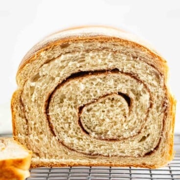 A close-up of a loaf of cinnamon swirl bread, sliced to show the spiral pattern inside, resting on a wire cooling rack with a light background.
