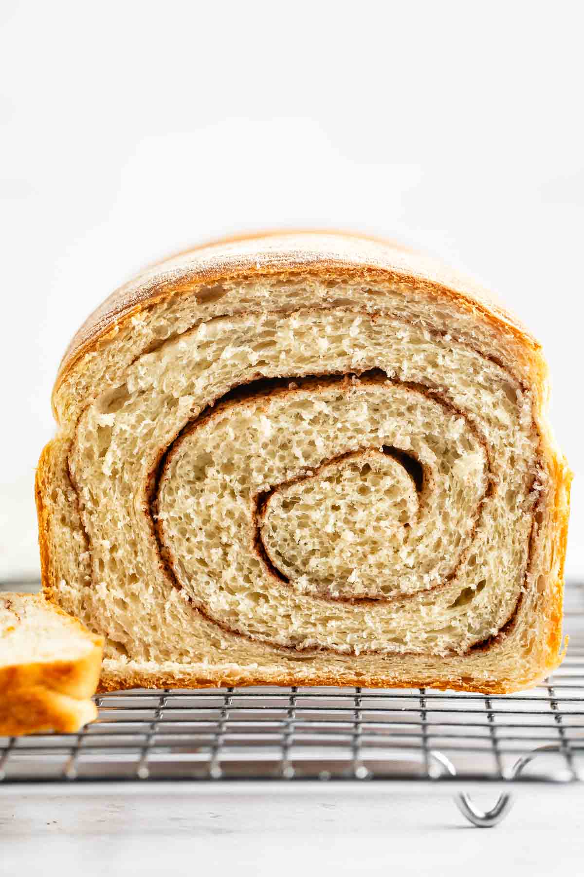 A close-up of a loaf of cinnamon swirl bread, sliced to show the spiral pattern inside, resting on a wire cooling rack with a light background.