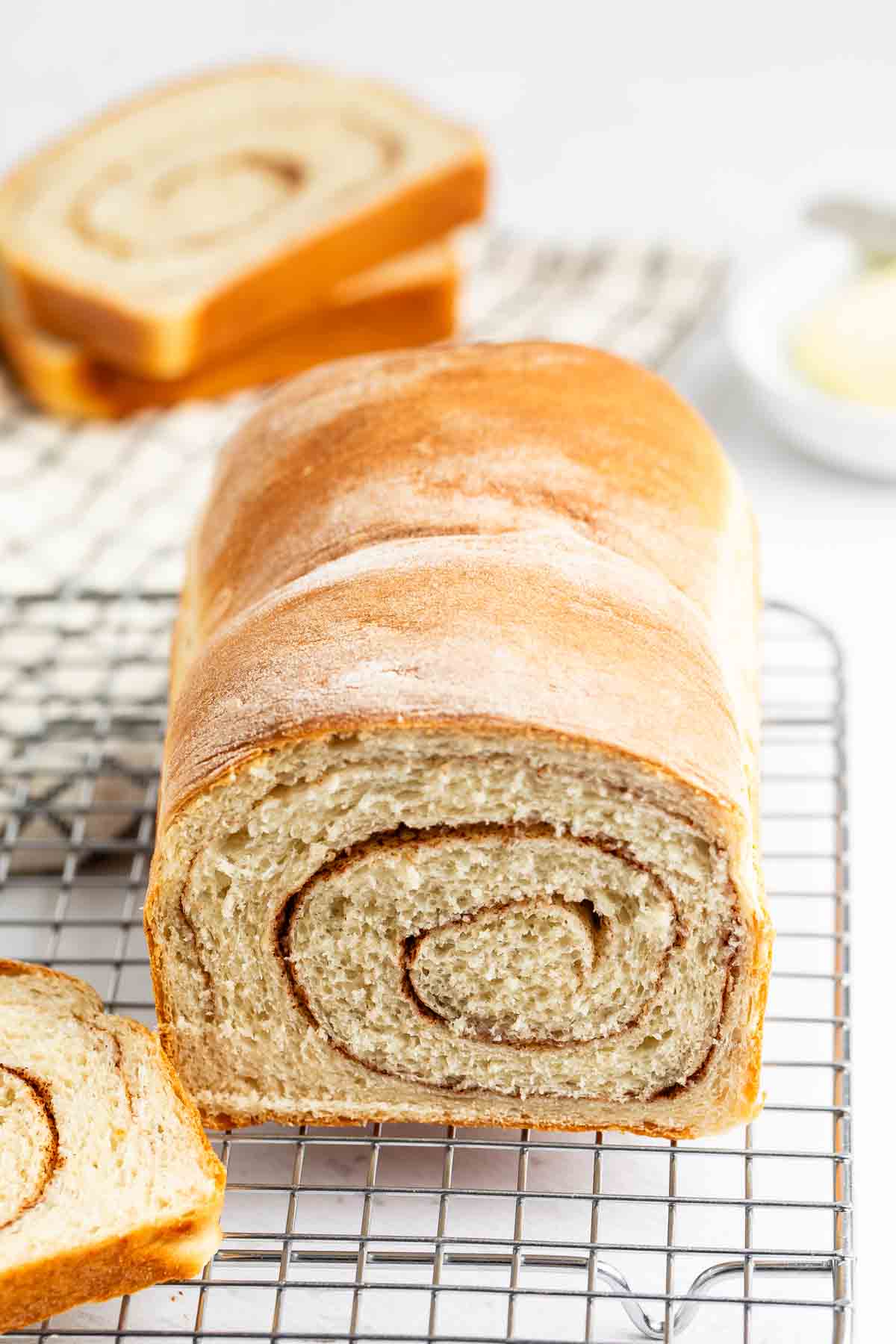 A loaf of cinnamon swirl bread rests on a cooling rack, with one slice cut and two more slices in the background. The bread has a golden-brown crust and a visible cinnamon spiral inside.