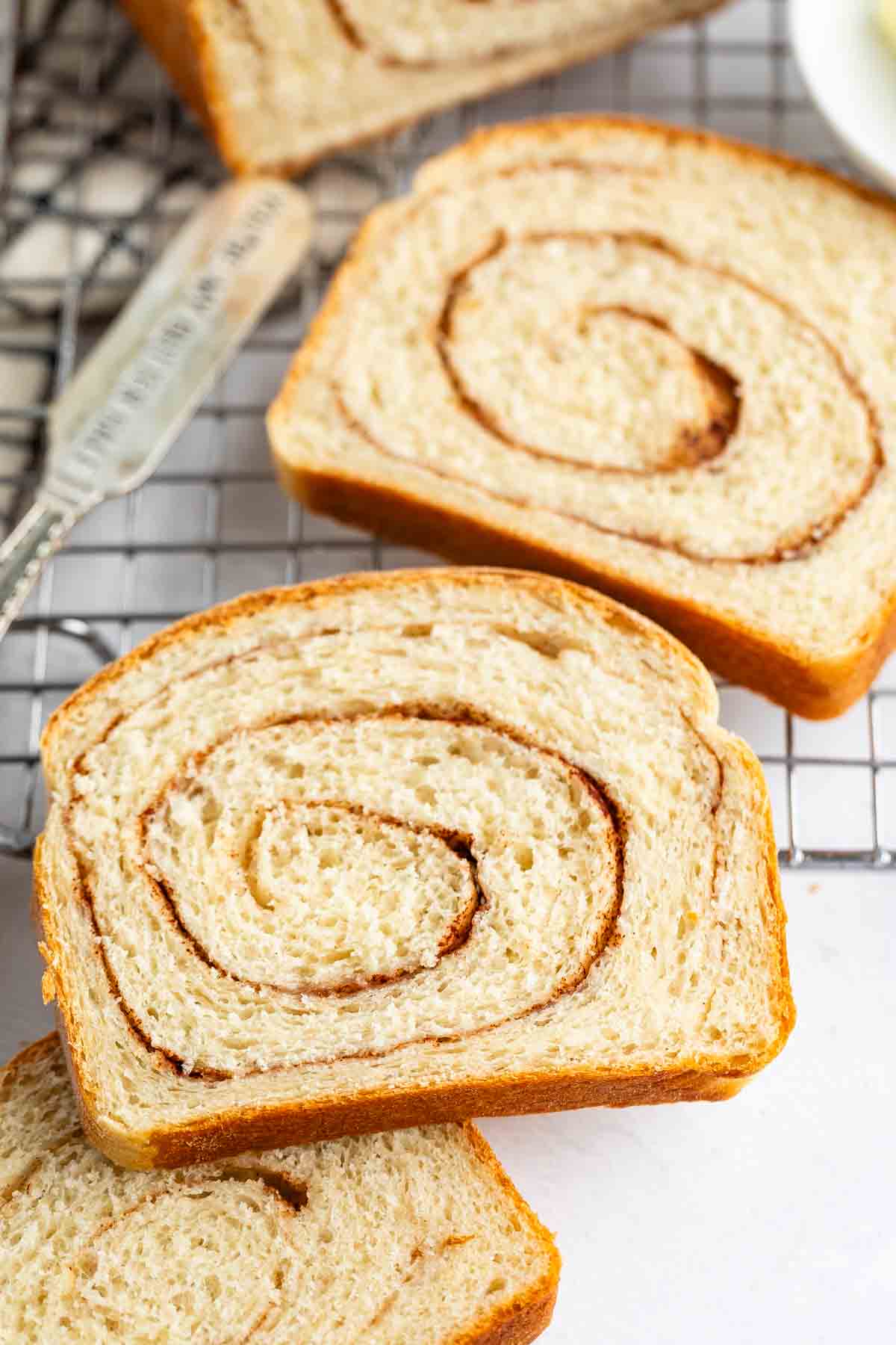 Slices of cinnamon swirl bread are placed on a cooling rack, showing a spiral pattern of cinnamon inside the soft, golden-brown bread. A butter knife is visible in the background.