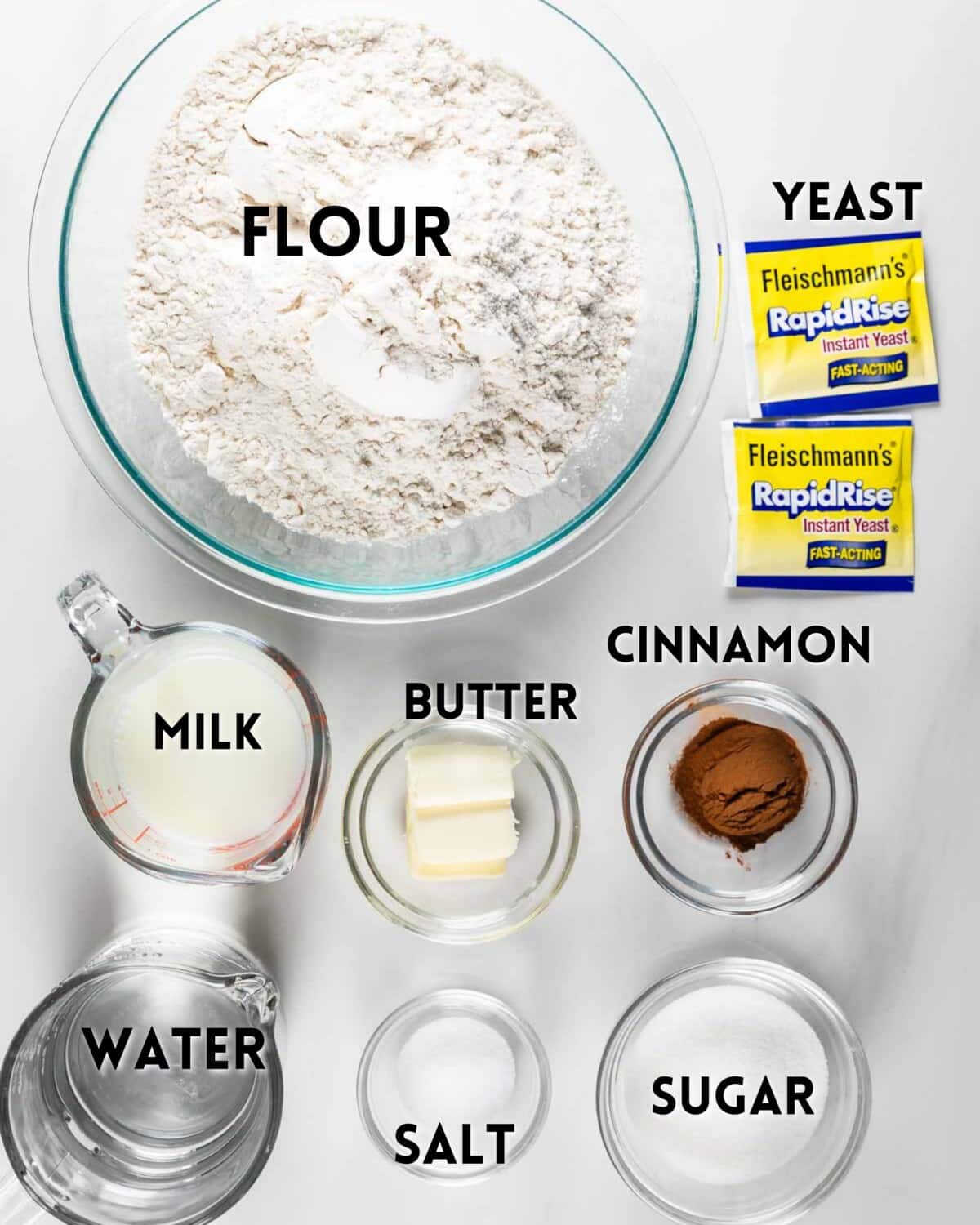Various baking ingredients labeled: a bowl of flour, packets of yeast, a cup of milk, a bowl of butter, a bowl of cinnamon, a cup of water, a bowl of salt, and a bowl of sugar arranged on a white surface.