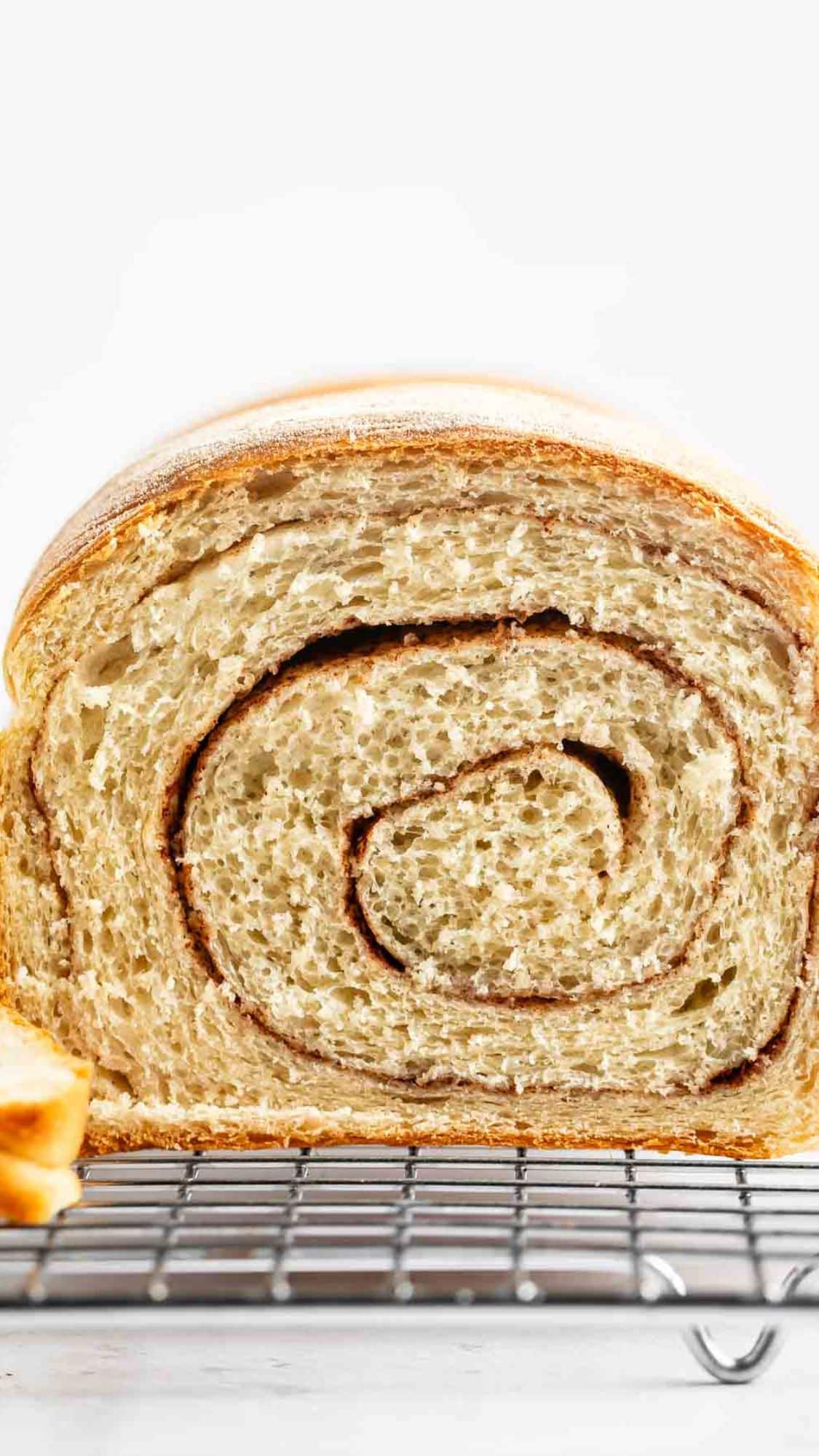 A close-up of a sliced homemade cinnamon swirl bread loaf, showing its soft texture and spiral pattern, resting on a cooling rack against a white background.