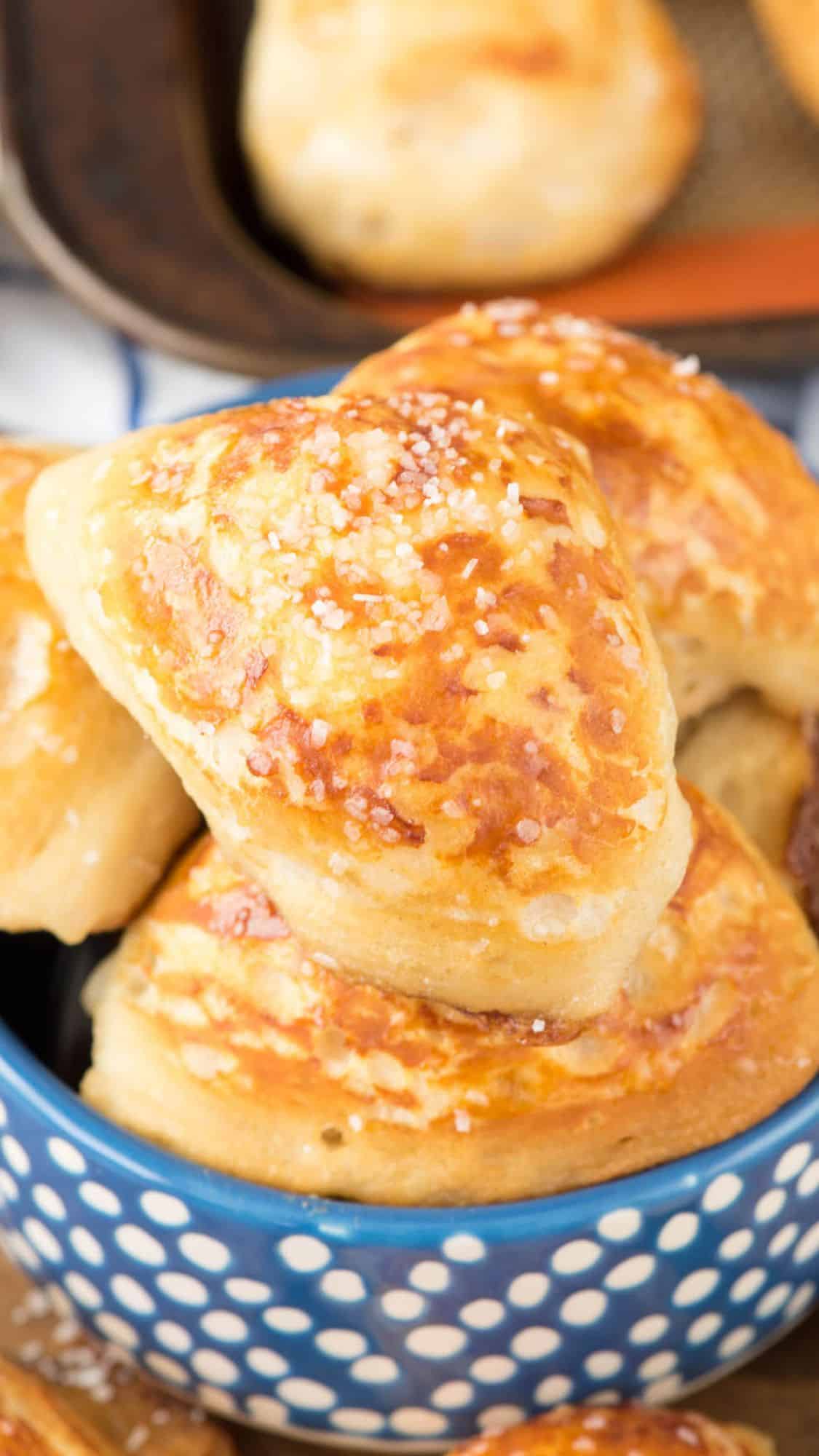 Golden-brown soft pretzel bites topped with coarse salt are stacked in a blue bowl with white polka dots, with more pretzel bites visible in the background on a baking tray.