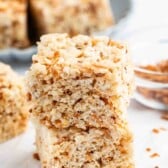 Two rice crispy treat bars stacked on top of each other, with a plate of more bars and a small glass bowl of toasted bits in the background on a white surface.