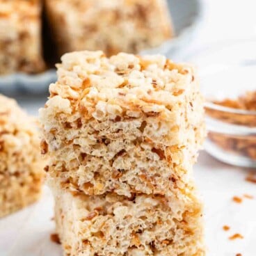 Two rice crispy treat bars stacked on top of each other, with a plate of more bars and a small glass bowl of toasted bits in the background on a white surface.
