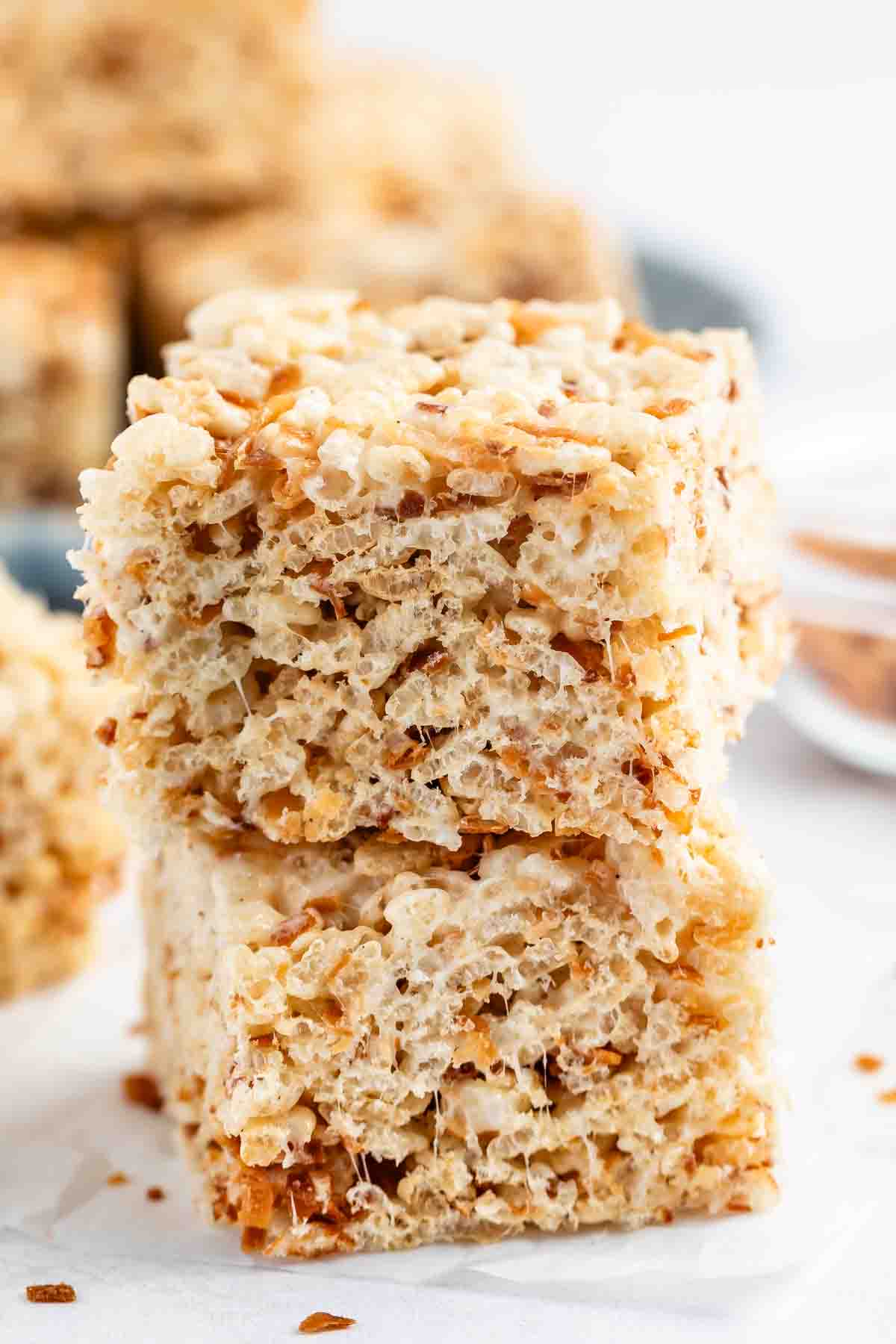 Two golden, gooey rice crispy treats stacked on top of each other, showing their crispy, airy texture and sticky marshmallow binding, with more treats blurred in the background.