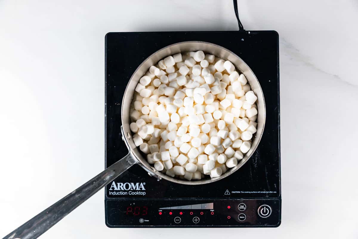 A pot filled with mini marshmallows sits on an Aroma induction cooktop, with a metal spoon resting in the mixture, seen from above on a white surface.