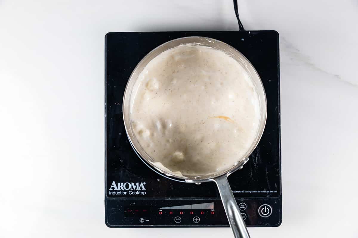 A saucepan filled with thick, creamy white sauce is heating on an Aroma induction cooktop, seen from above against a white background.