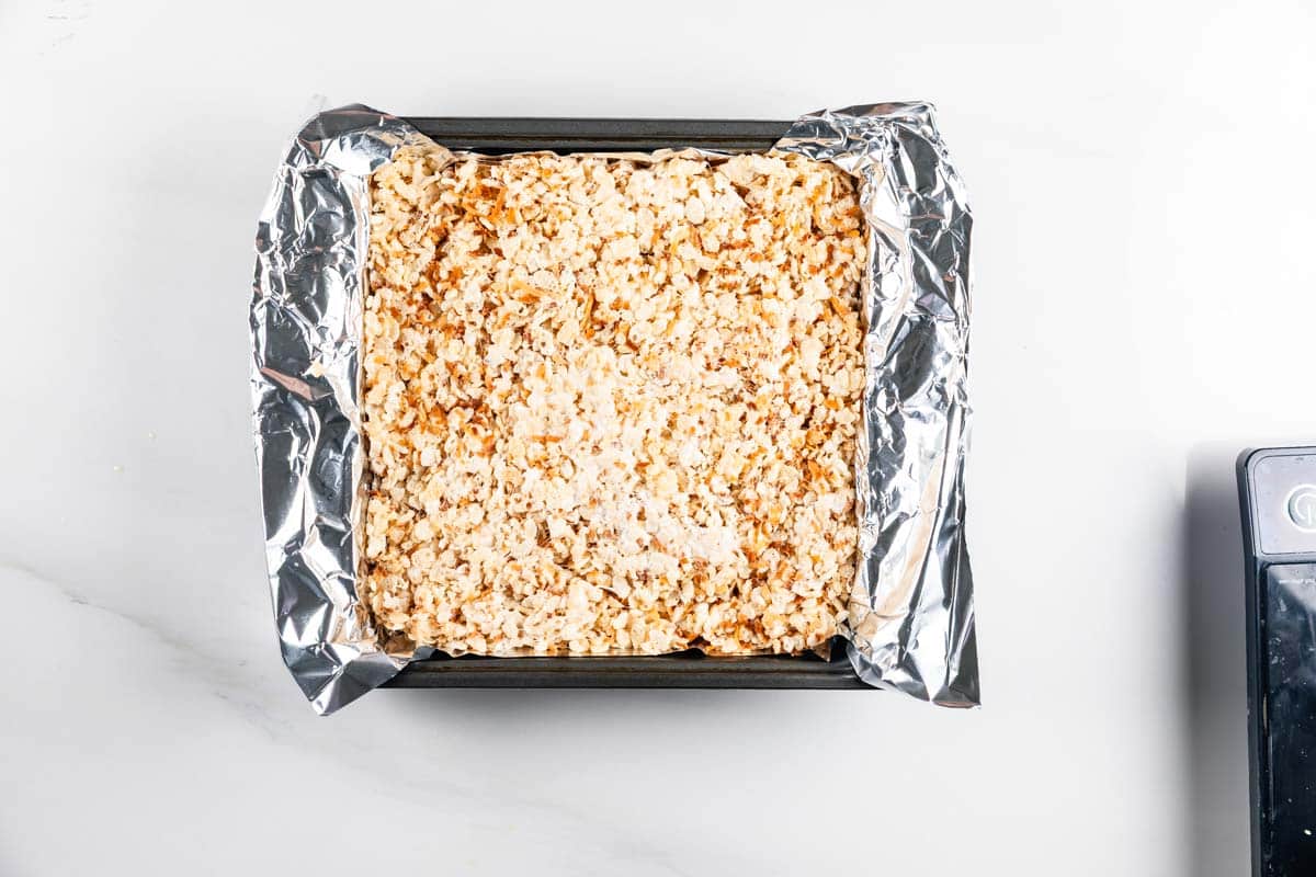 A square baking pan lined with aluminum foil is filled with a pressed mixture of rice cereal treats, viewed from above on a white surface.