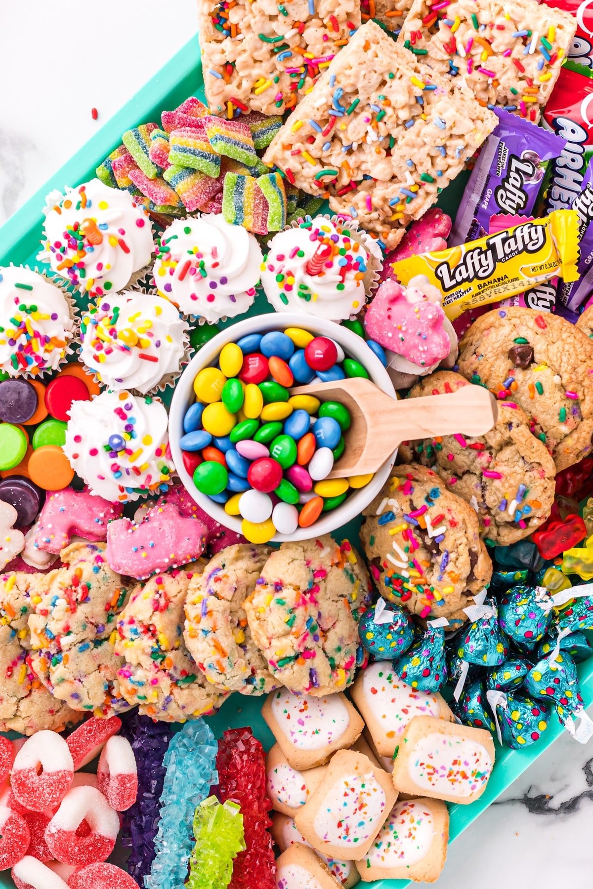 A colorful platter filled with cookies, cupcakes, candies, marshmallows, rainbow gummies, Rice Krispies treats, and a bowl of M&Ms, all topped with sprinkles, displayed on a turquoise tray.
