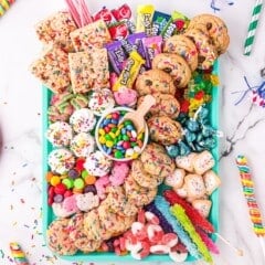 A teal tray filled with assorted colorful cookies, candies, gummies, chocolate, and sweets, surrounded by party decorations, confetti, and plates on a white marble surface.