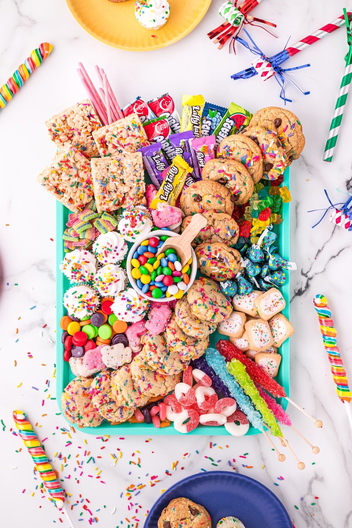A teal tray filled with assorted colorful cookies, candies, gummies, chocolate, and sweets, surrounded by party decorations, confetti, and plates on a white marble surface.