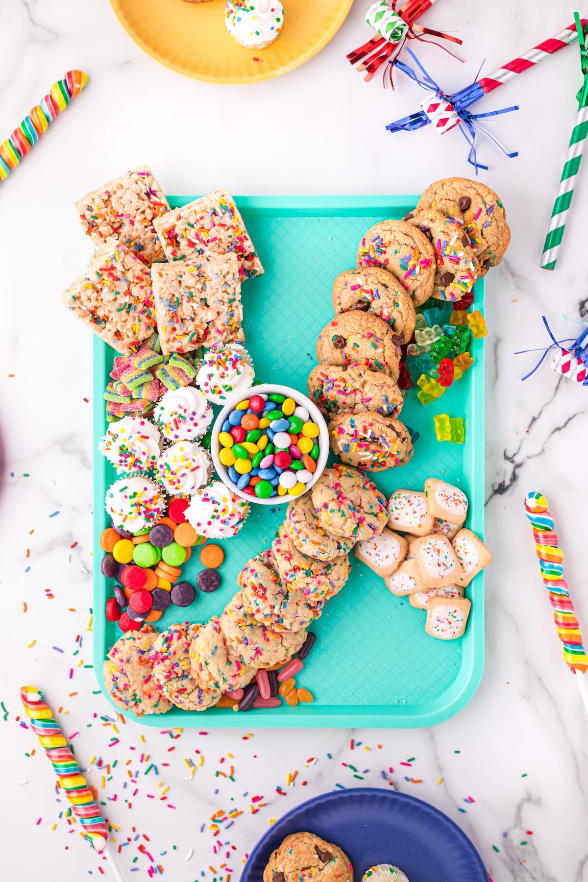 A turquoise tray holds assorted colorful cookies, candies, and sprinkles. There are chocolate chip cookies, frosted cookies, and sweets. Party horns, plates, and scattered sprinkles decorate the marble surface around the tray.