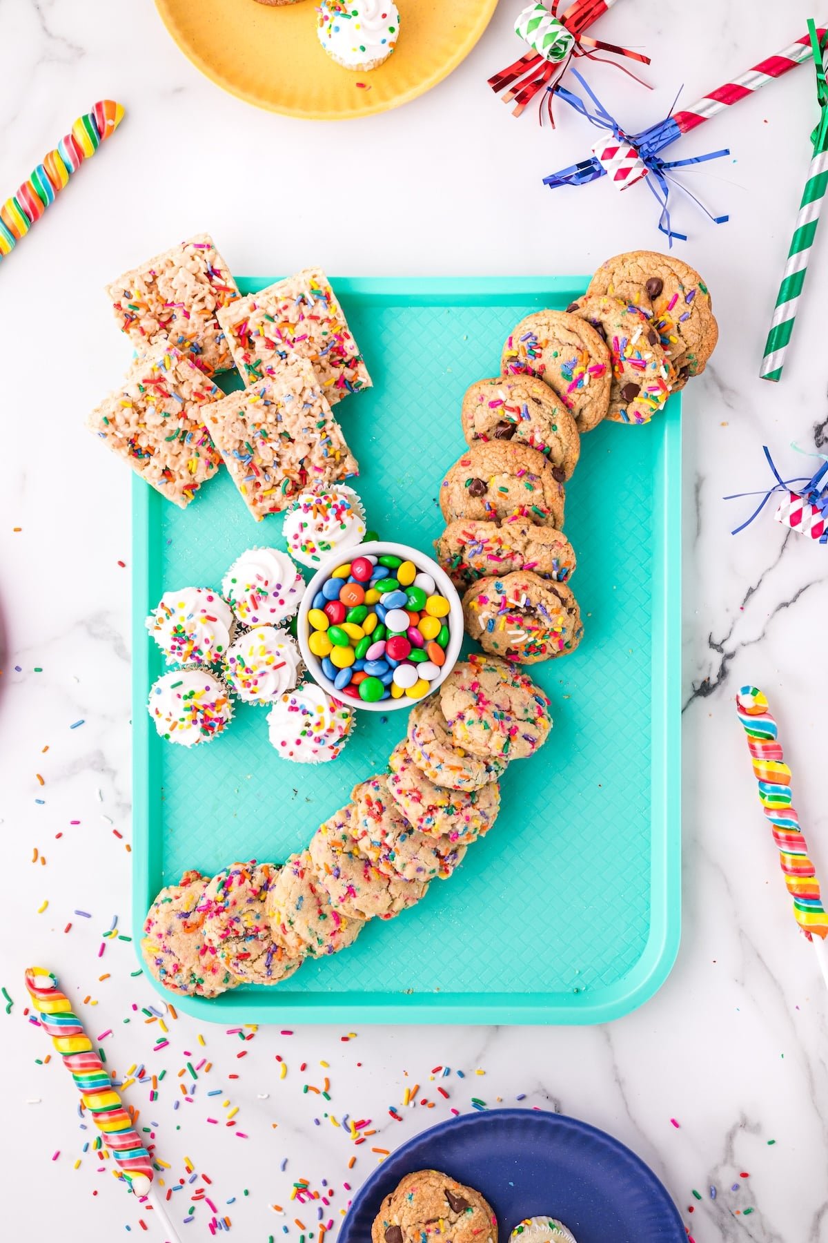A turquoise tray displays cookies, rice crispy treats, and sprinkle-topped treats arranged in the shape of a 7, with a small bowl of colorful candy in the center. Party favors and plates surround the tray on a white surface.