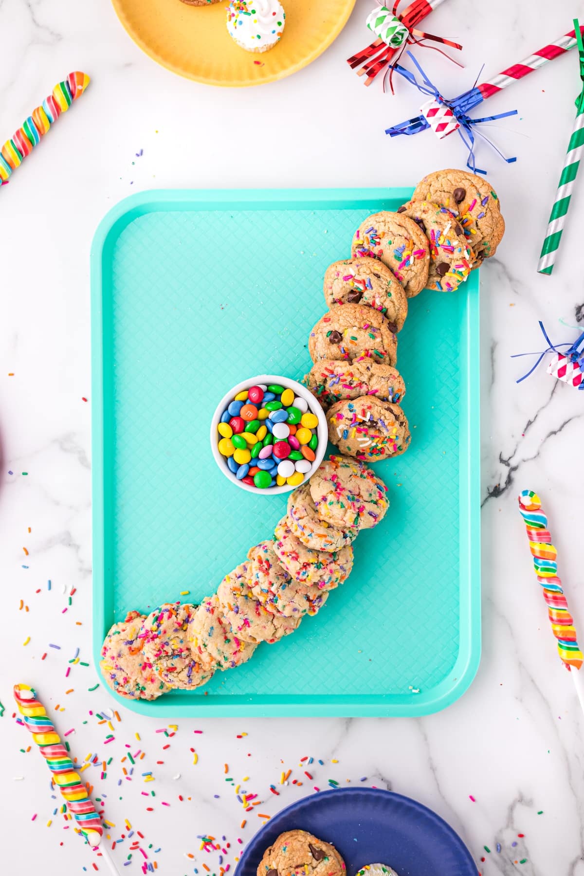 A turquoise tray holds a bowl of colorful candies surrounded by a curved row of sprinkle cookies. Party decorations, confetti, and plates with desserts are scattered around on a marble surface.