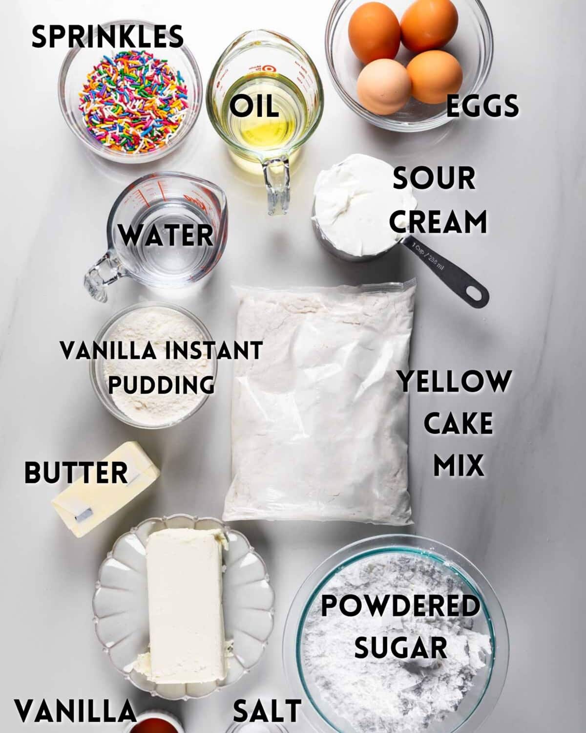 Overhead view of cake ingredients on a white surface, including sprinkles, oil, eggs, sour cream, water, vanilla instant pudding, yellow cake mix, butter, powdered sugar, vanilla, and salt, each labeled.