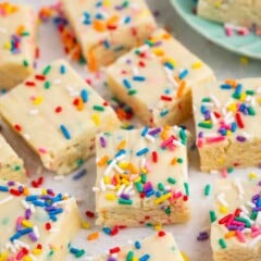 Several frosted, sprinkle-topped sugar cookie bars are arranged on parchment paper, with colorful rainbow sprinkles scattered around. Some bars are stacked on a green plate in the background.