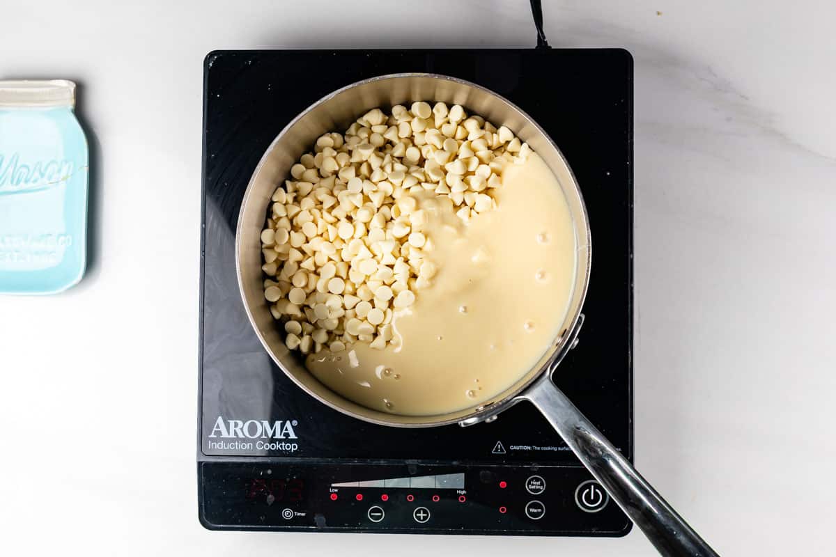 A saucepan with white chocolate chips and sweetened condensed milk sits on an Aroma induction cooktop, ready to be melted.