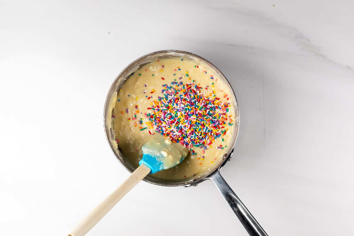 A saucepan filled with cake batter topped with colorful rainbow sprinkles, being stirred with a blue spatula on a white surface.