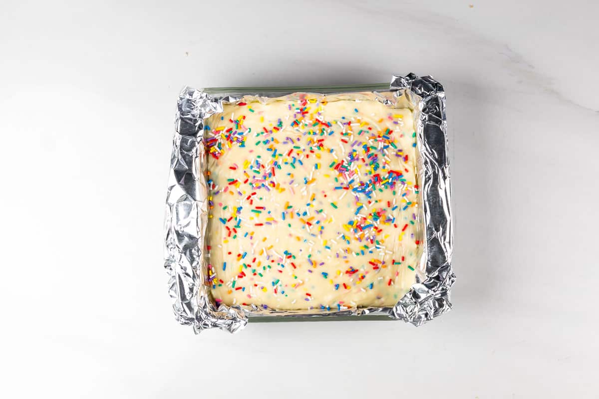 A square baking pan lined with foil holds a cake topped with white icing and colorful rainbow sprinkles, viewed from above on a white surface.
