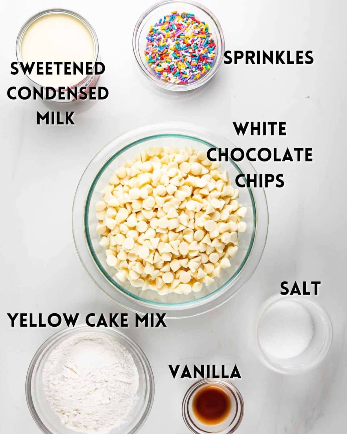 Top-down view of labeled ingredients in bowls for a recipe: sweetened condensed milk, rainbow sprinkles, white chocolate chips, yellow cake mix, salt, and vanilla, all arranged on a white surface.
