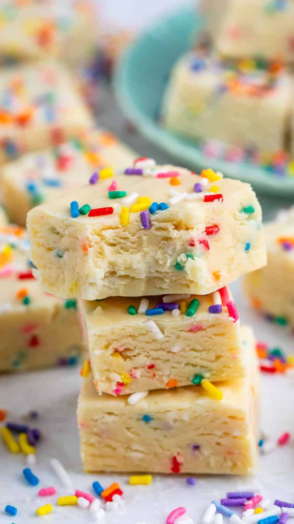 Three square pieces of white fudge with colorful rainbow sprinkles are stacked on top of each other, with more sprinkled fudge pieces and loose sprinkles in the background.
