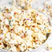 A bowl of popcorn covered in colorful rainbow sprinkles, with extra popcorn and sprinkles scattered on a white surface around the bowl.