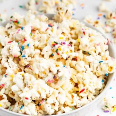 A bowl of popcorn covered in colorful rainbow sprinkles, with extra popcorn and sprinkles scattered on a white surface around the bowl.