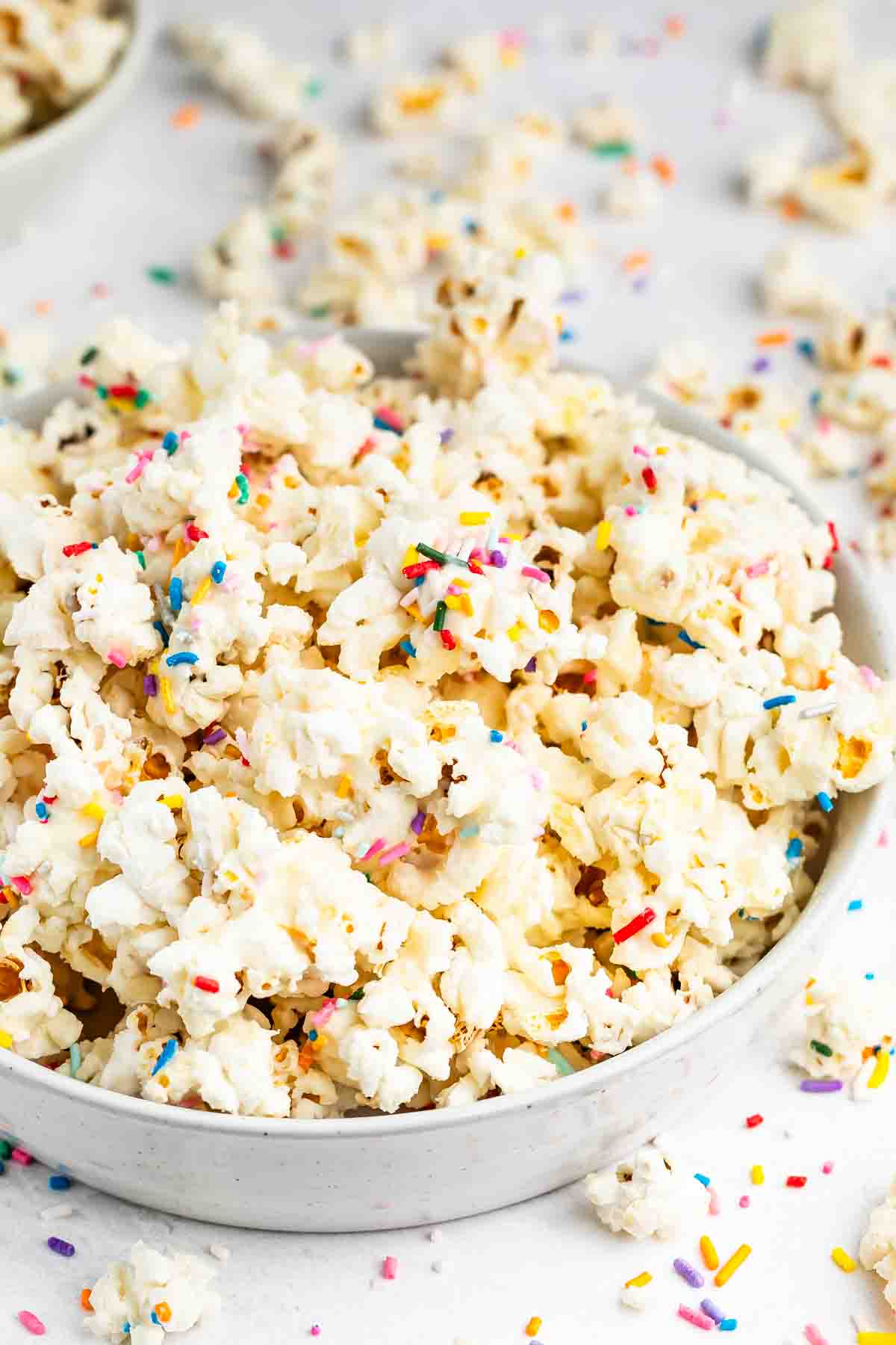 A bowl of popcorn covered in colorful rainbow sprinkles, with extra popcorn and sprinkles scattered on a white surface around the bowl.